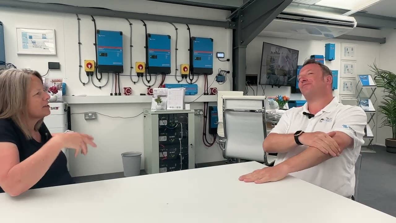 Victron inverter/şarj cihazı duvarının önündeki masada konuşan iki kişi — video karesi